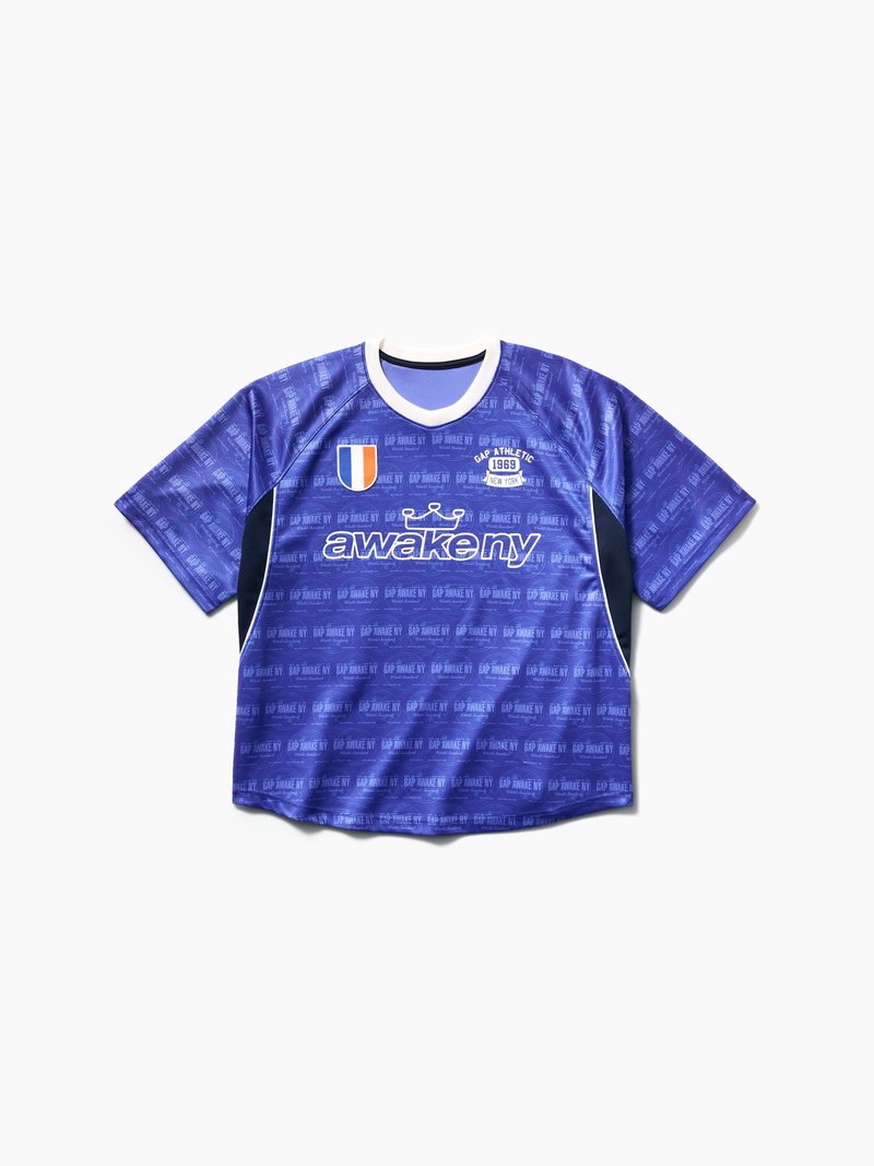 Blaues kurzärmeliges Sporttrikot mit weißem Kragen, schwarzen Seitenpartien, dem Schriftzug „awake ny“ und athletischen Aufnähern auf der Brust.