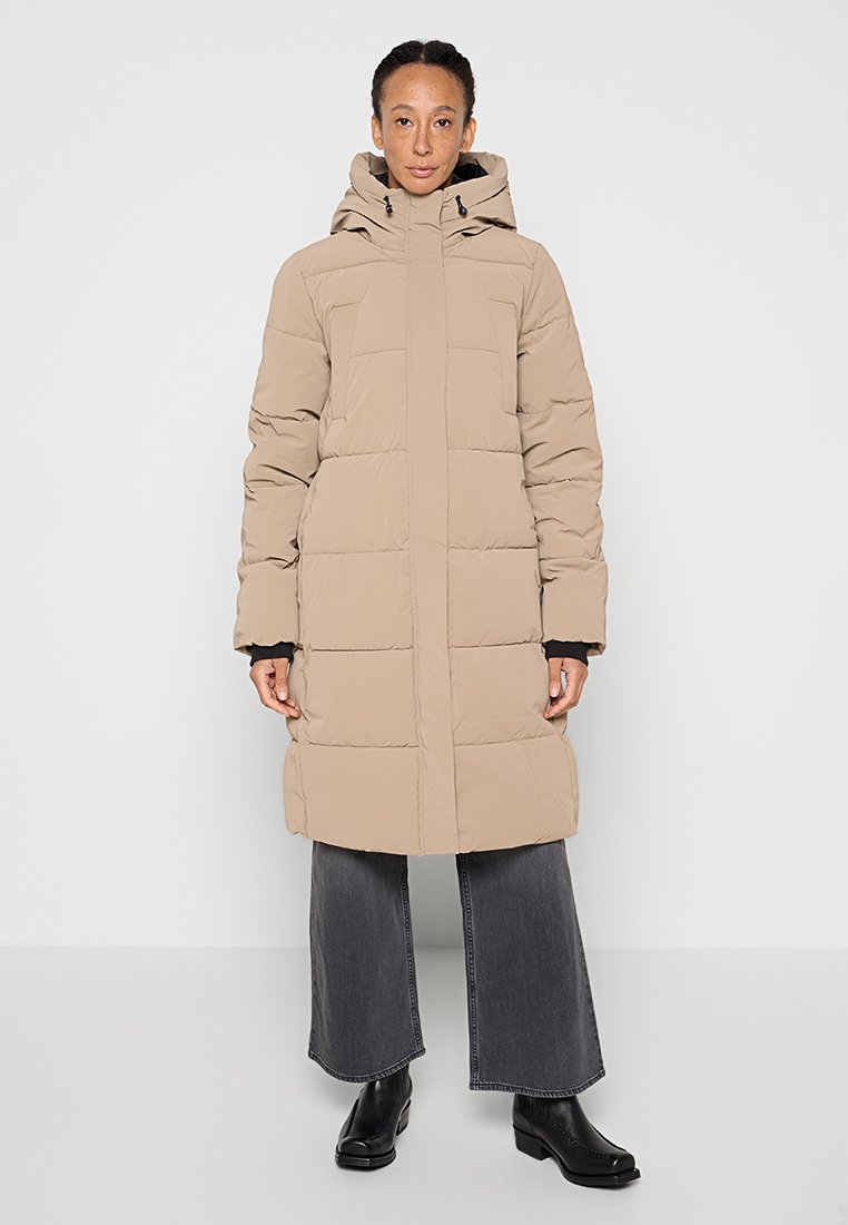 Frau trägt eine beige lange Steppjacke mit Kapuze, schwarze weit geschnittene Jeans und schwarze Stiefeletten, steht vor einem einfarbigen Hintergrund.