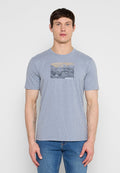 Mann trägt ein hellgraues kurzärmeliges T-Shirt mit abstraktem Bergmotiv und "MEXX"-Logo, kombiniert mit blauer Jeans, steht vor weißem Hintergrund.