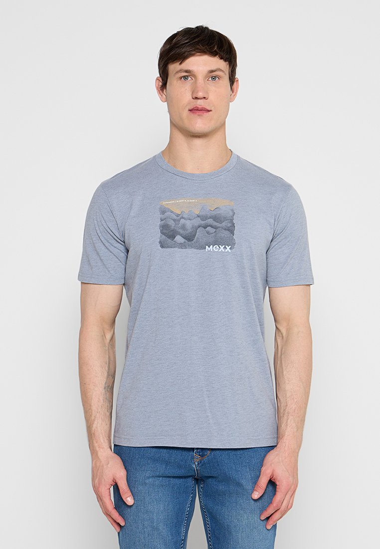 Mann trägt ein hellgraues kurzärmeliges T-Shirt mit abstraktem Bergmotiv und "MEXX"-Logo, kombiniert mit blauer Jeans, steht vor weißem Hintergrund.
