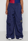 Marineblaue Cargo-Hosen aus glänzendem, leichtem Stoff, mit Seitentaschen und einem dekorativen gelben Emblem am Oberschenkel.