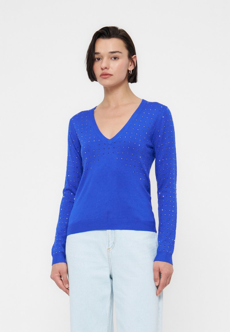 Frau trägt einen blauen Langarm-V-Ausschnitt-Pullover mit kleinen Nieten und hellblauen Jeans, steht vor einem schlichten weißen Hintergrund.