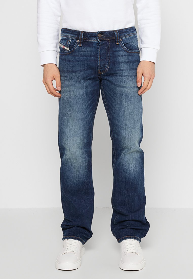 Dunkelblaue Jeans aus Denim mit geradem Bein Schnitt, die leichte Verblassungen aufweisen, mit einem Knopfverschluss und fünf Taschen, getragen mit weißen Turnschuhen.