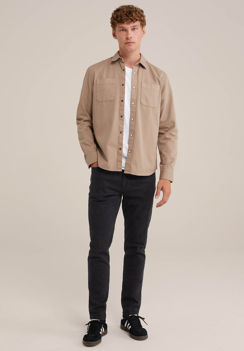 Beige Hemd mit Knopfverschluss und zwei Brusttaschen, über einem weißen T-Shirt gestylt, kombiniert mit schwarzen Hosen und schwarzen Sneakern. Einfarbiger Hintergrund.