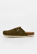 Einzelner olivgrüner Wildleder-Slideschuh mit einem silbernen Schnallenriemen und einem Kork-Fußbett mit weißer Sohle vor schlichtem Hintergrund.