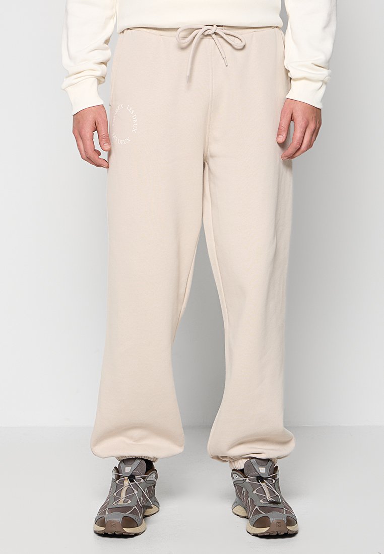 Mann in beigen Sweatpants mit Kordelzugbund und elastischen Bündchen, kombiniert mit grau-braunen Sneakers, steht vor einem schlichten Hintergrund.