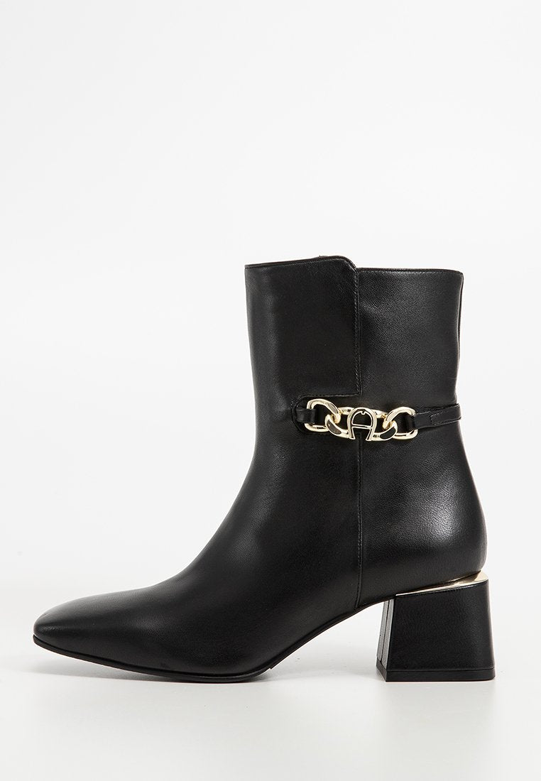 Schwarze Lederknöchelstiefel mit spitzem Zeh und Blockabsatz. Mit einem goldenen Kettenakzent am Knöchel und einer glatten, matten Textur.