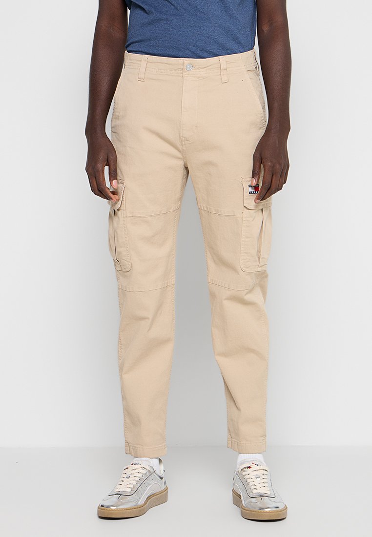Mann trägt beige Cargo-Hosen mit Seitentaschen, ein blaues Hemd, weiße Socken und weiße Sneaker mit Gummisohlen, steht vor hellem Hintergrund.