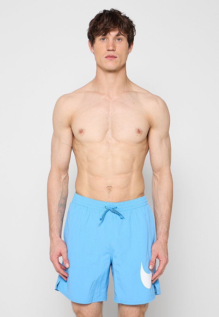 Oberkörperfreier Mann mit kurzem dunklem Haar trägt hellblaue Badeshorts mit weißem Logo und steht vor schlichtem hellgrauem Hintergrund.