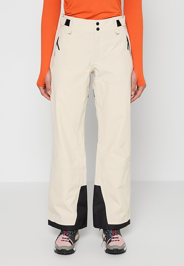 Beige Weitbein-Hose mit schwarzen Einsätzen an den Bündchen, seitlichen Reißverschlusstaschen, getragen mit orangefarbenem Langarm-Top und mehrfarbigen Wanderschuhen.
