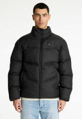Mann in einer schwarzen gepolsterten Jacke mit einem Reißverschluss auf der Vorderseite und hellblauen Jeans, der vor einem weißen Hintergrund steht.