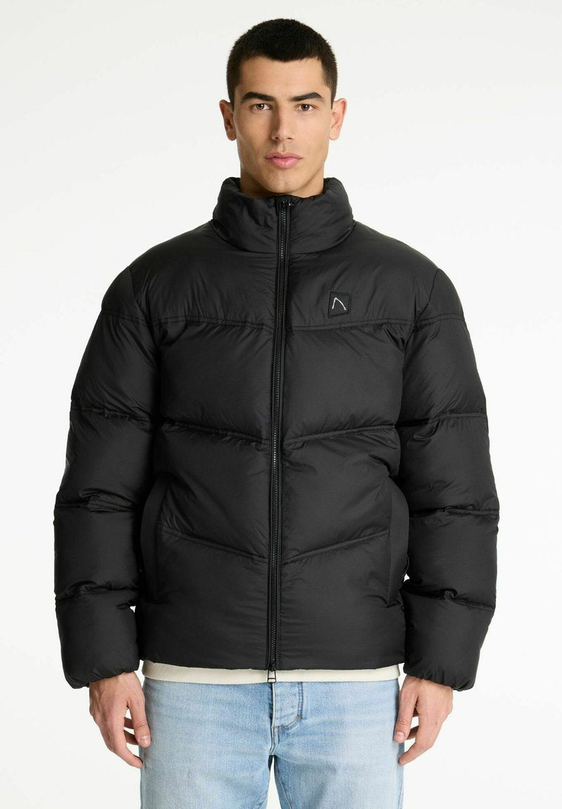 Mann in einer schwarzen gepolsterten Jacke mit einem Reißverschluss auf der Vorderseite und hellblauen Jeans, der vor einem weißen Hintergrund steht.