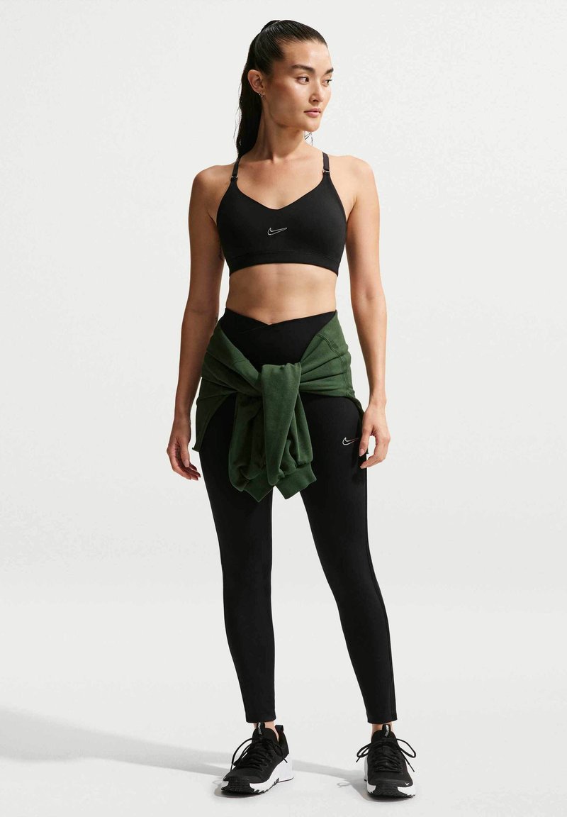 Frau trägt schwarzen Sport-BH, schwarze Leggings, schwarz-weiße Turnschuhe und hat einen grünen Pullover um die Taille gebunden, steht vor weißem Hintergrund.