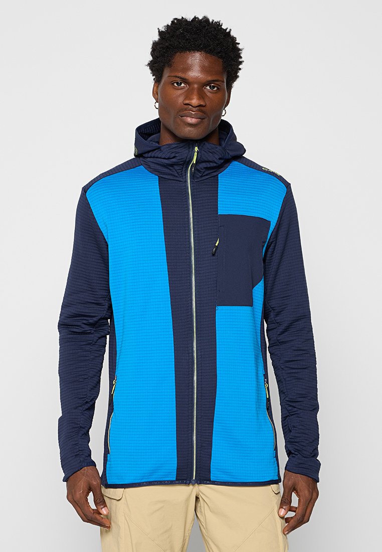 Mann in einer blauen und marineblauen Zip-Up-Hoodie-Jacke mit Brusttasche und beigen Hosen, stehend vor einem schlichten hellen Hintergrund.