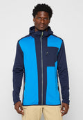 Mann in einer blauen und marineblauen Zip-Up-Hoodie-Jacke mit Brusttasche und beigen Hosen, stehend vor einem schlichten hellen Hintergrund.