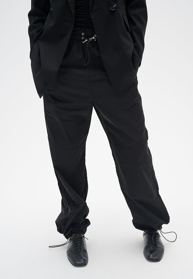 Schwarze, locker sitzende Hosen mit einem elastischen Bund und Kordelzug. Glatte Textur mit leicht glänzendem Finish, kombiniert mit schwarzen Geschäftsschuhen.