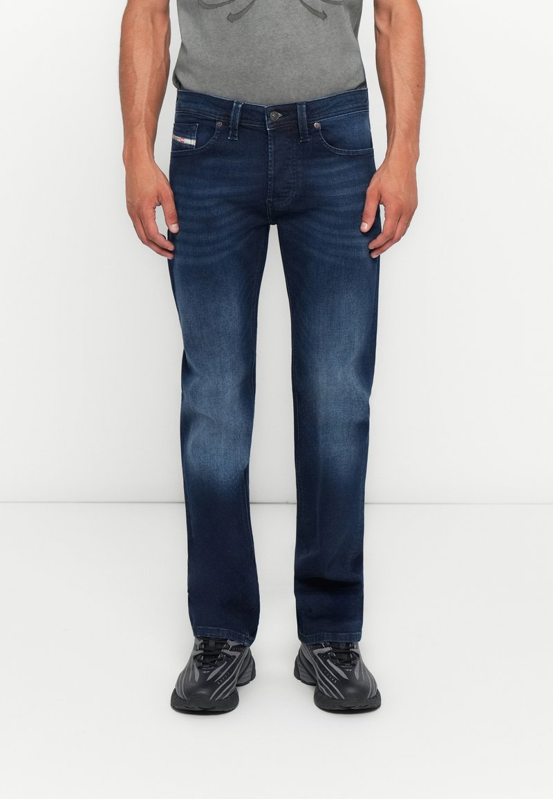 Mann in dunkelblauen lässigen Jeans, grauem T-Shirt und schwarzen Turnschuhen, der vor einem schlichten weißen Hintergrund steht.