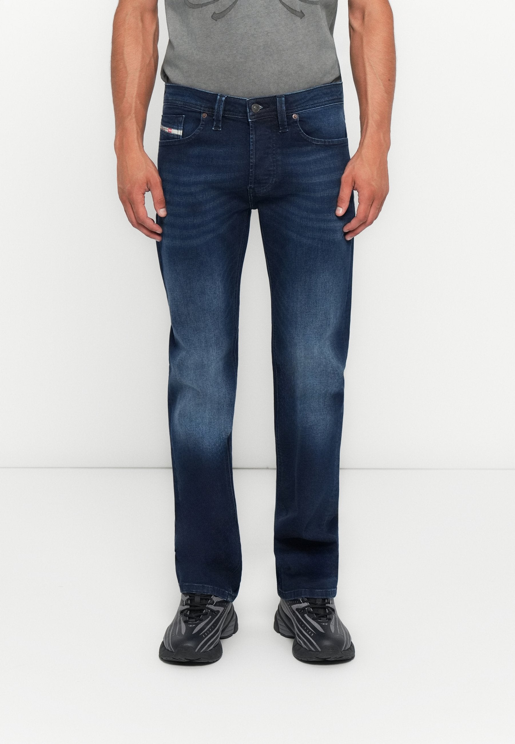 Mann in dunkelblauen lässigen Jeans, grauem T-Shirt und schwarzen Turnschuhen, der vor einem schlichten weißen Hintergrund steht.
