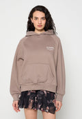 Frau mit lockigem dunklem Haar, trägt einen taupefarbenen AllSaints-Hoodie und einen schwarzen Blumenrock, steht vor einem schlichten hellen Hintergrund.
