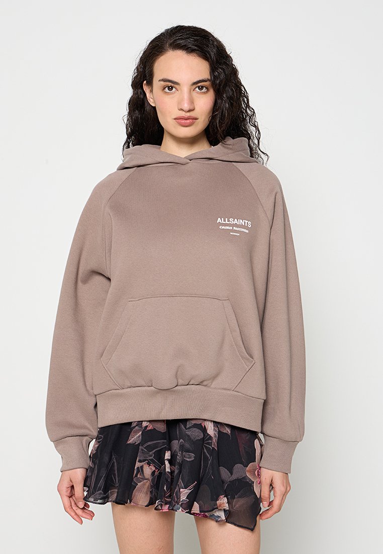 Frau mit lockigem dunklem Haar, trägt einen taupefarbenen AllSaints-Hoodie und einen schwarzen Blumenrock, steht vor einem schlichten hellen Hintergrund.