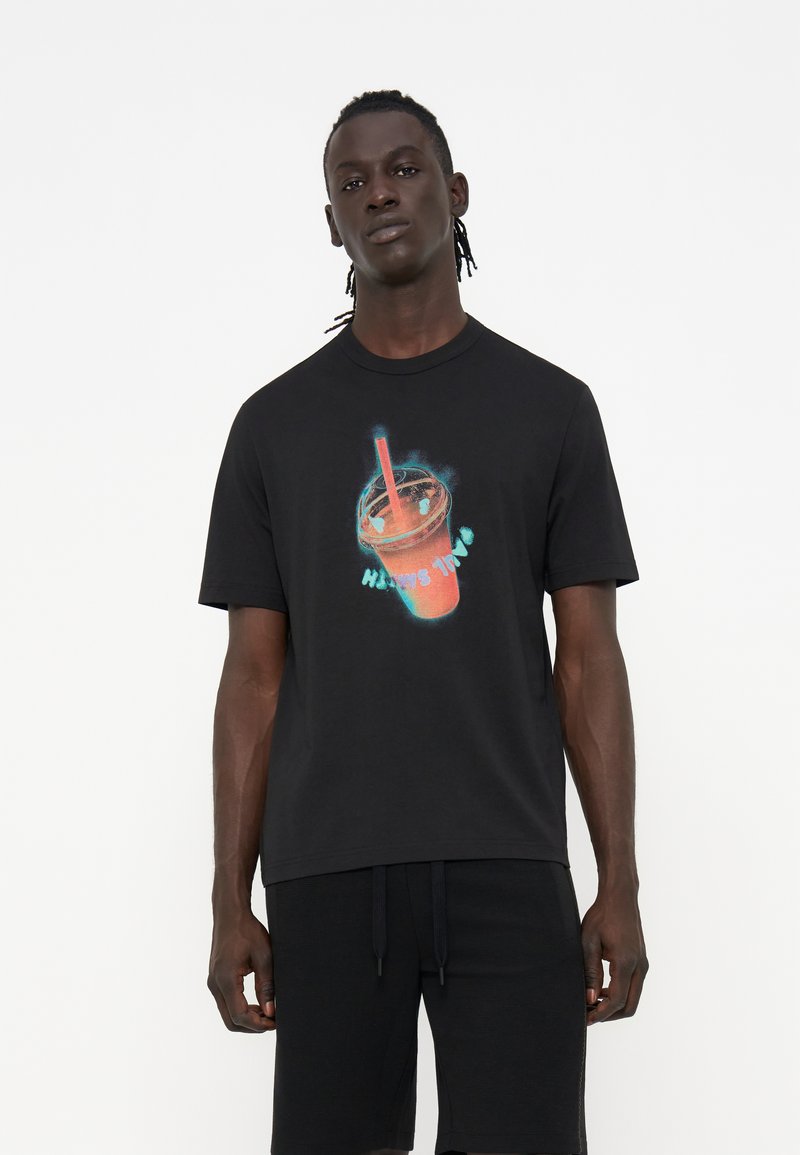 Junger Mann mit dunkler Haut und geflochtenem Haar, der ein schwarzes T-Shirt mit einer leuchtenden Smoothie-Becher-Grafik und dem Text „Happy Time“ trägt.