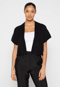 Schwarze gestrickte, kurzärmelige Cropped-Cardigan mit geripptem Muster und offenem Design. Kombiniert mit einem weißen Oberteil und schwarzen Hosen.