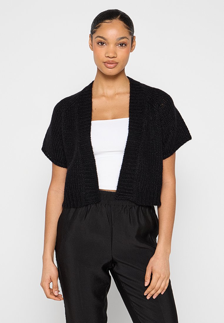Schwarze gestrickte, kurzärmelige Cropped-Cardigan mit geripptem Muster und offenem Design. Kombiniert mit einem weißen Oberteil und schwarzen Hosen.