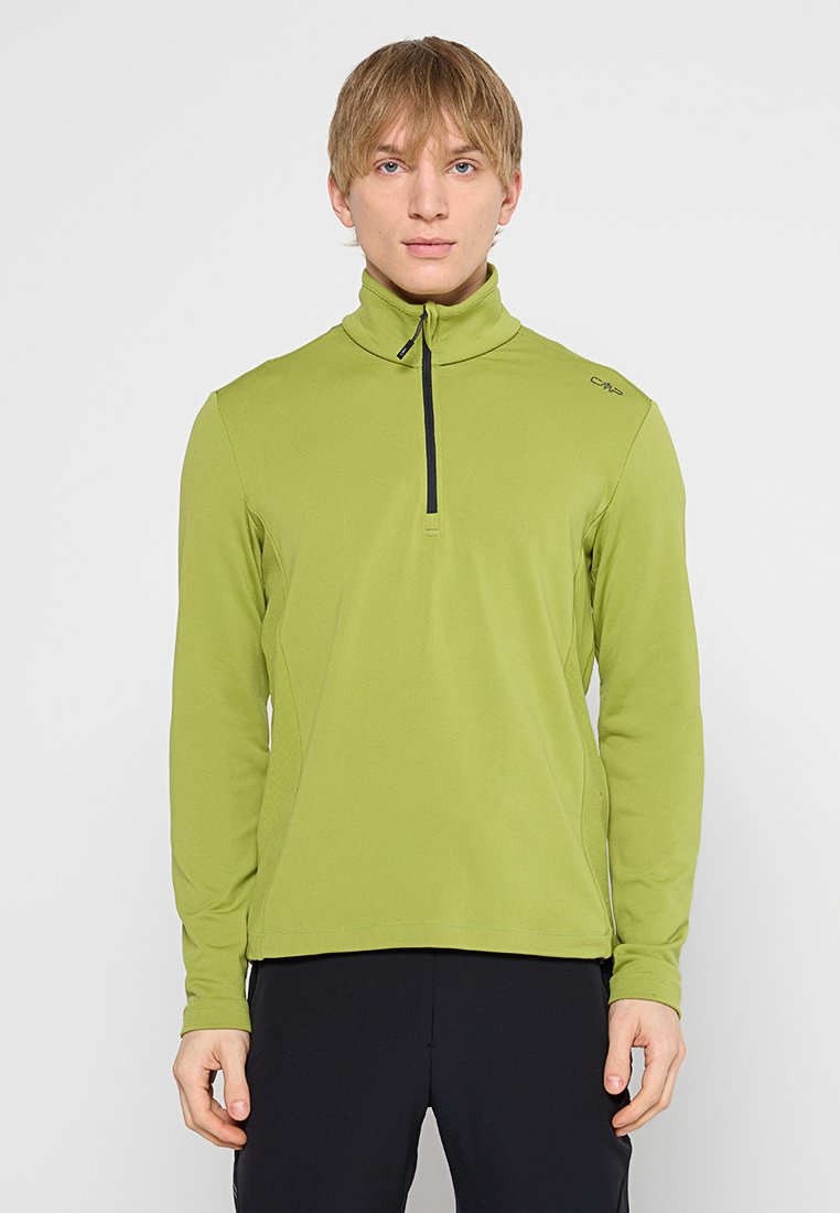 Junger Mann, der einen limettengrünen Langarmpullover mit einem Viertelreißverschluss trägt und vor einem schlichten hellgrauen Hintergrund steht.