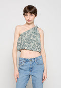 Frau trägt ein grünes und weiß gemustertes Crop-Top mit einem Schulterriemen, kombiniert mit hellblauen, hoch taillierten Jeans, steht vor einem einfarbigen Hintergrund.
