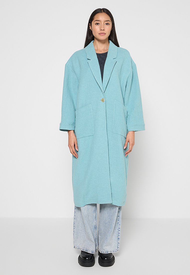 Langer, hellblauer Mantel aus strukturiertem Stoff, mit Fronttaschen, einer Knopfschließe und oversized Passform, kombiniert mit weit geschnittenen Jeans und schwarzen Schuhen.