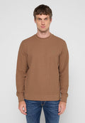 Mann in einem braunen Langarm-Waffelpullover mit dezentem Logo auf der Brust und blauen Jeans, der vor einem schlichten weißen Hintergrund steht.