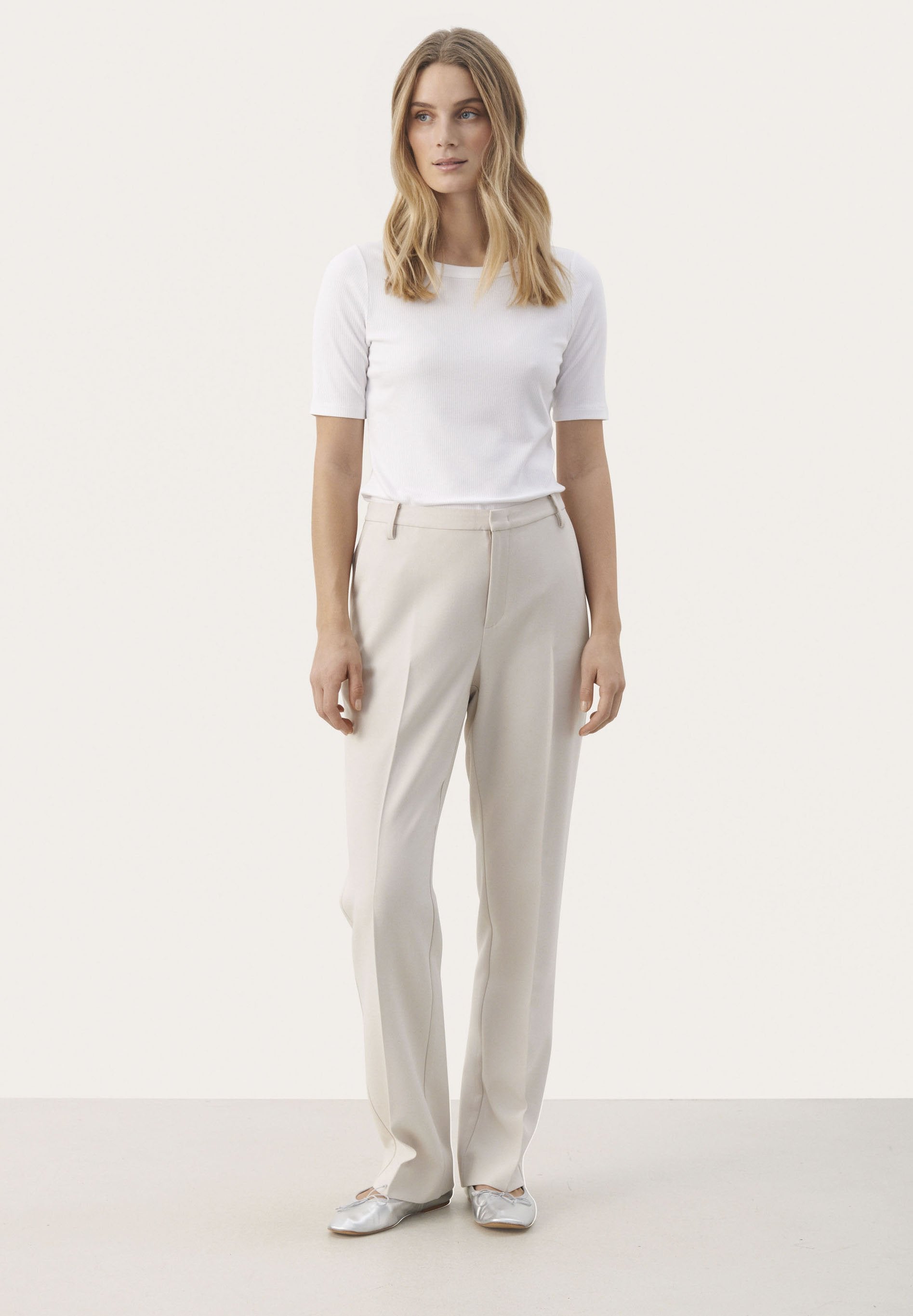 Beige Hose aus strukturiertem Stoff, gerade geschnitten, mit Taschen auf der Vorderseite und hohem Bund. Kombiniert mit einem figurbetonten weißen Kurzarmoberteil.