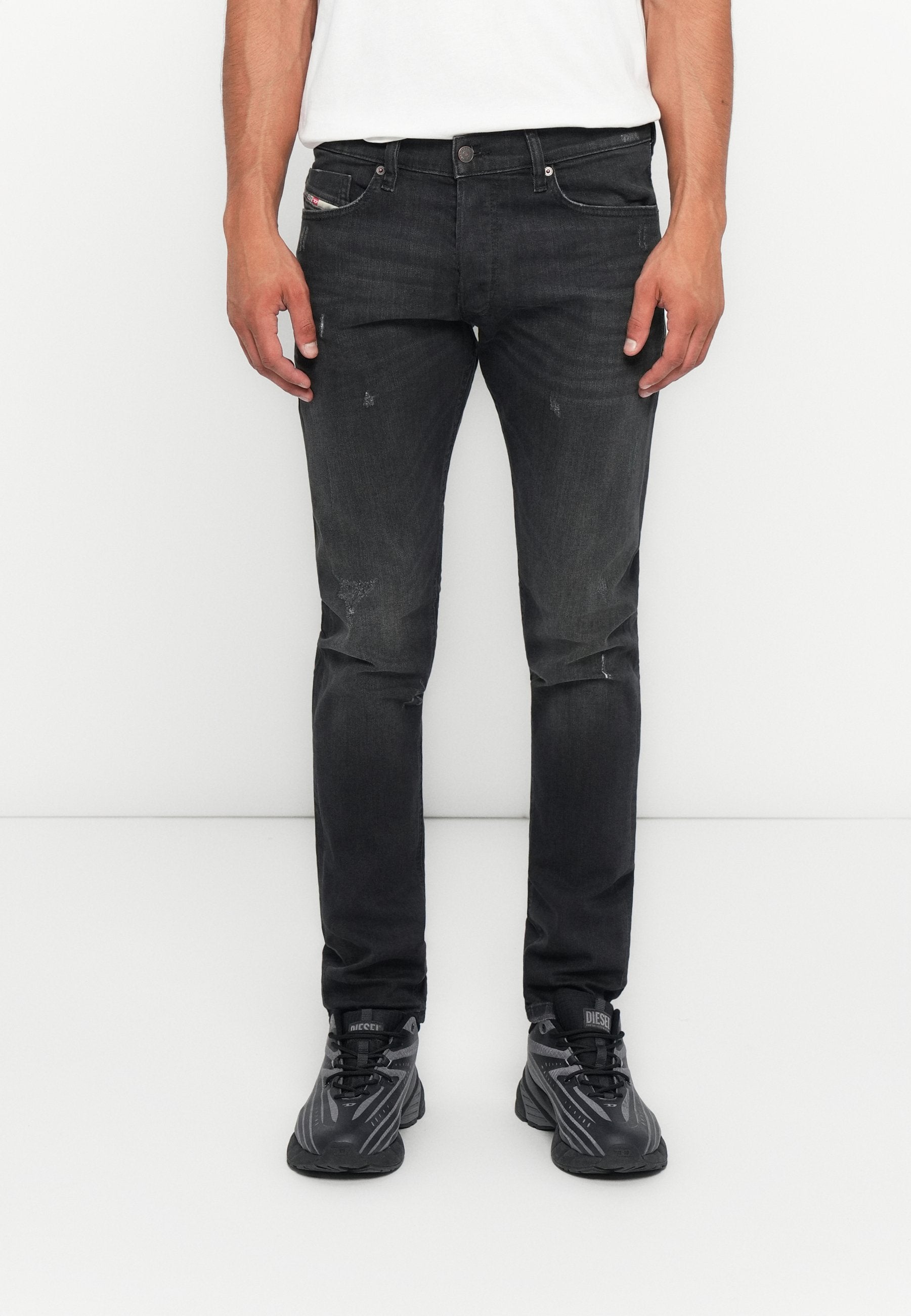 Mann in eng geschnittenen, ausgebleichten schwarzen Jeans mit dezenten Used-Effekten und schwarzen Diesel-Sneakern, vor einem schlichten weißen Hintergrund stehend.