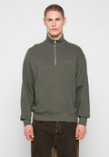 Mann trägt einen olivgrünen Pullover mit halbem Reißverschluss und dunkelbraune Hose, steht vor einem einfarbigen hellgrauen Hintergrund.