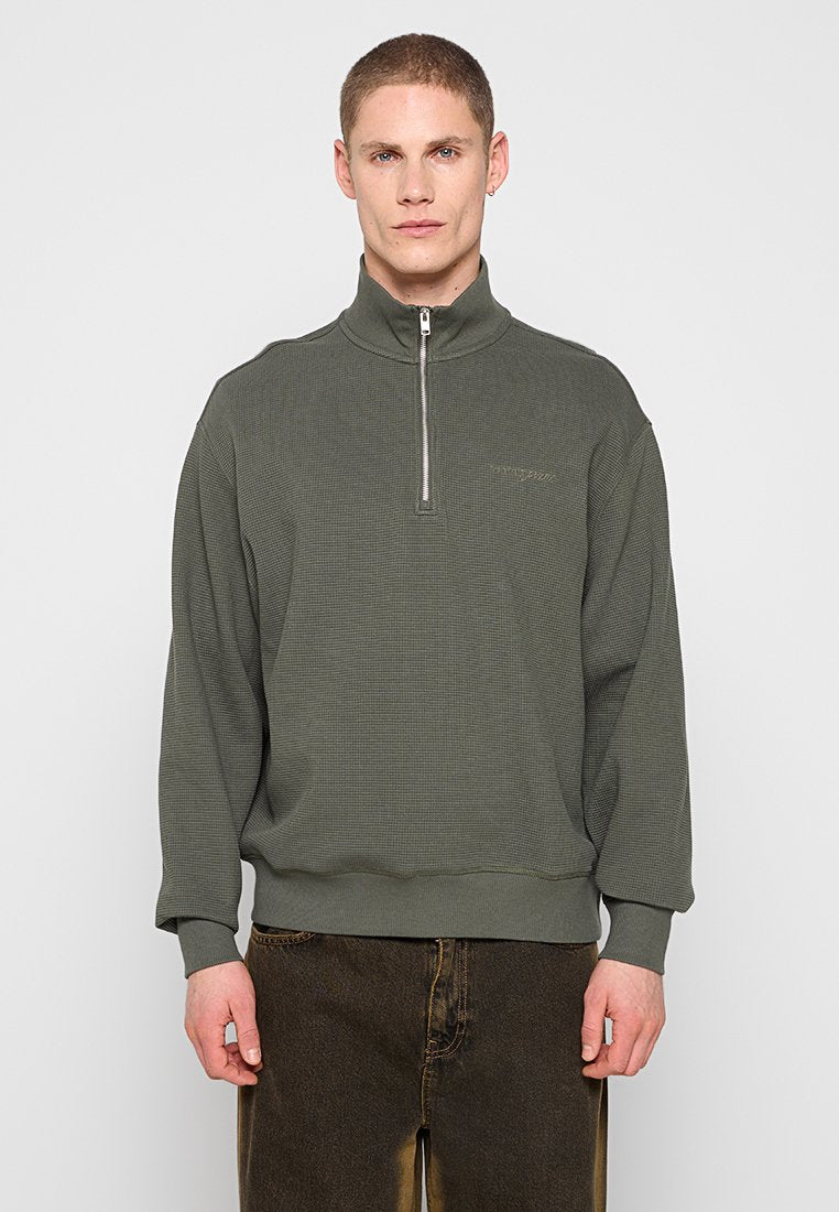 Mann trägt einen olivgrünen Pullover mit halbem Reißverschluss und dunkelbraune Hose, steht vor einem einfarbigen hellgrauen Hintergrund.