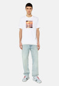 Weißes T-Shirt mit kurzen Ärmeln und einem quadratischen Grafikdesign, das einen Sonnenuntergang über Wasser zeigt, kombiniert mit hellblauen Jeans und silbern gemusterten Schuhen.