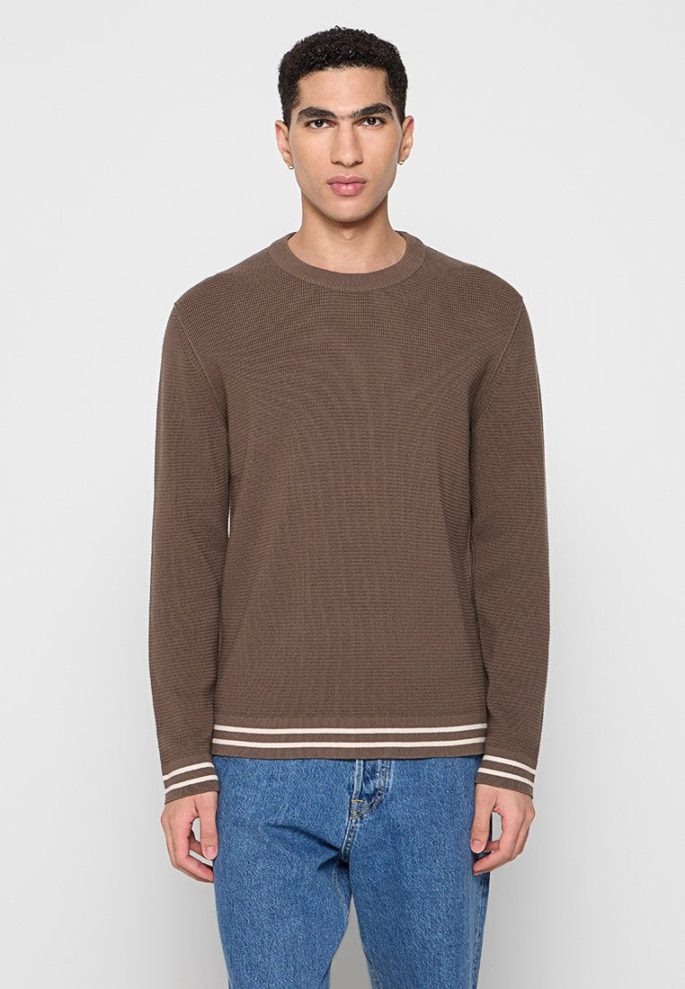 Brauner gerippter Pullover mit Rundhalsausschnitt, der weiße horizontale Streifen an den Bündchen und am Saum aufweist. weiche Textur, schmaler Schnitt. Darunter tragen Sie eine Jeans.
