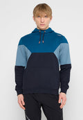 Junger Mann mit glattem blonden Haar, der einen blau-schwarz gefärbten Hoodie mit einer Fronttasche und schwarze Hosen vor einem einfarbigen Hintergrund trägt.