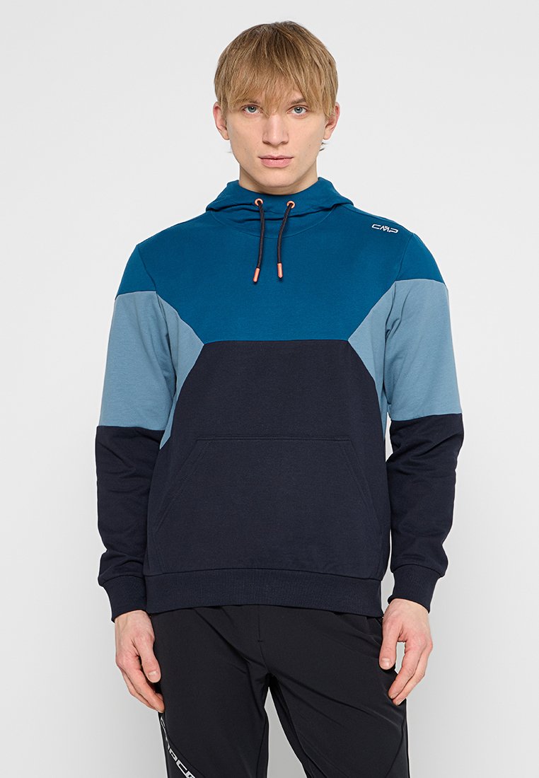 Junger Mann mit glattem blonden Haar, der einen blau-schwarz gefärbten Hoodie mit einer Fronttasche und schwarze Hosen vor einem einfarbigen Hintergrund trägt.