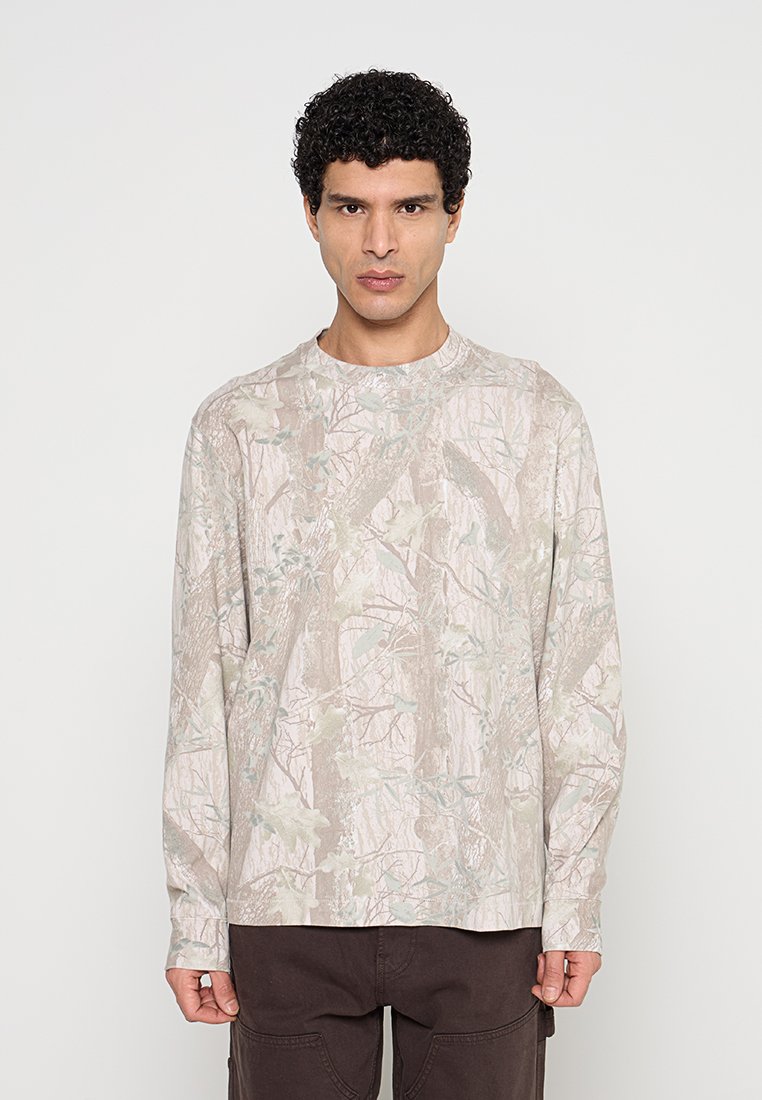 Mann mit kurzen, lockigen schwarzen Haaren, der ein langärmliges beige Hemd mit Tarnmuster und dunkelbraune Hose trägt, steht vor einem einfarbigen Hintergrund.