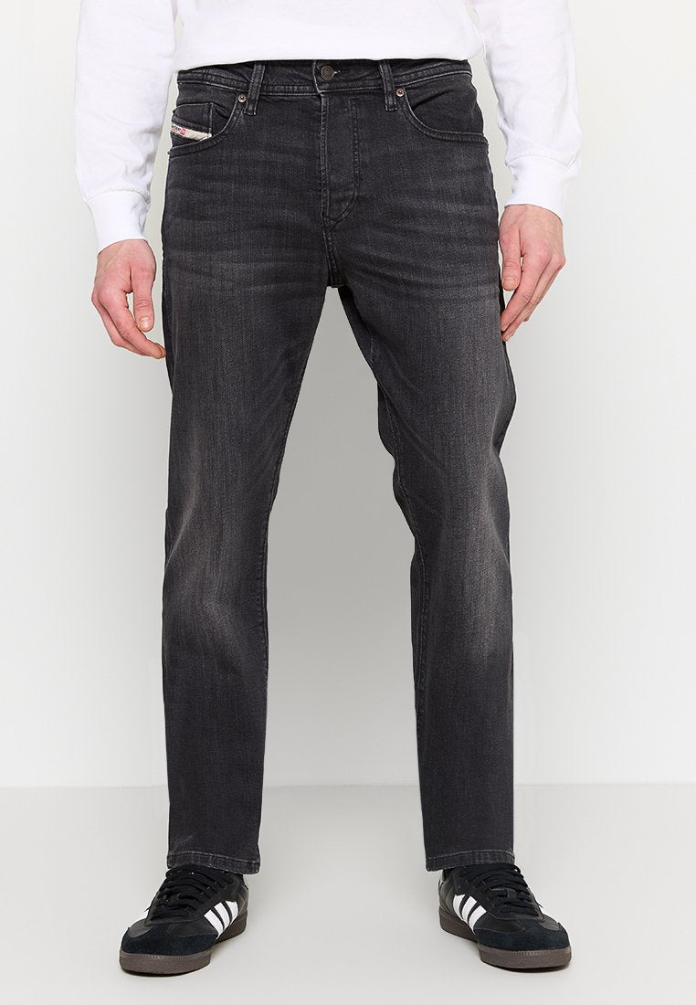 Dunkelgraue Jeans aus Denim mit geradem Schnitt, Fünf-Taschen-Design, subtilen Verfärbungen und metallischen Details. Getragen mit schwarzen Sneakers.