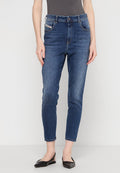 Frau, die schmal geschnittene, blaue Jeans mit vorderen und Münz- pockets trägt, kombiniert mit schwarzen spitzen Flats, steht vor einem einfarbigen Hintergrund.