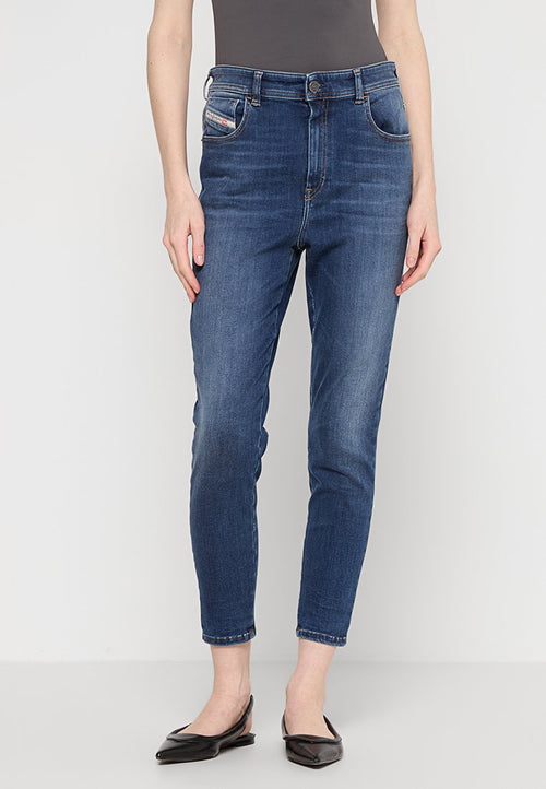 Frau, die schmal geschnittene, blaue Jeans mit vorderen und Münz- pockets trägt, kombiniert mit schwarzen spitzen Flats, steht vor einem einfarbigen Hintergrund.