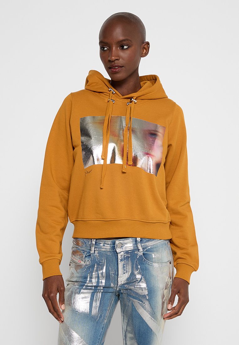 Senfgelb Hoodie mit glänzendem silbernen Grafikdruck, Zugbandkapuze und cropped Design. Kombiniert mit hellblauen, distressed Jeans.