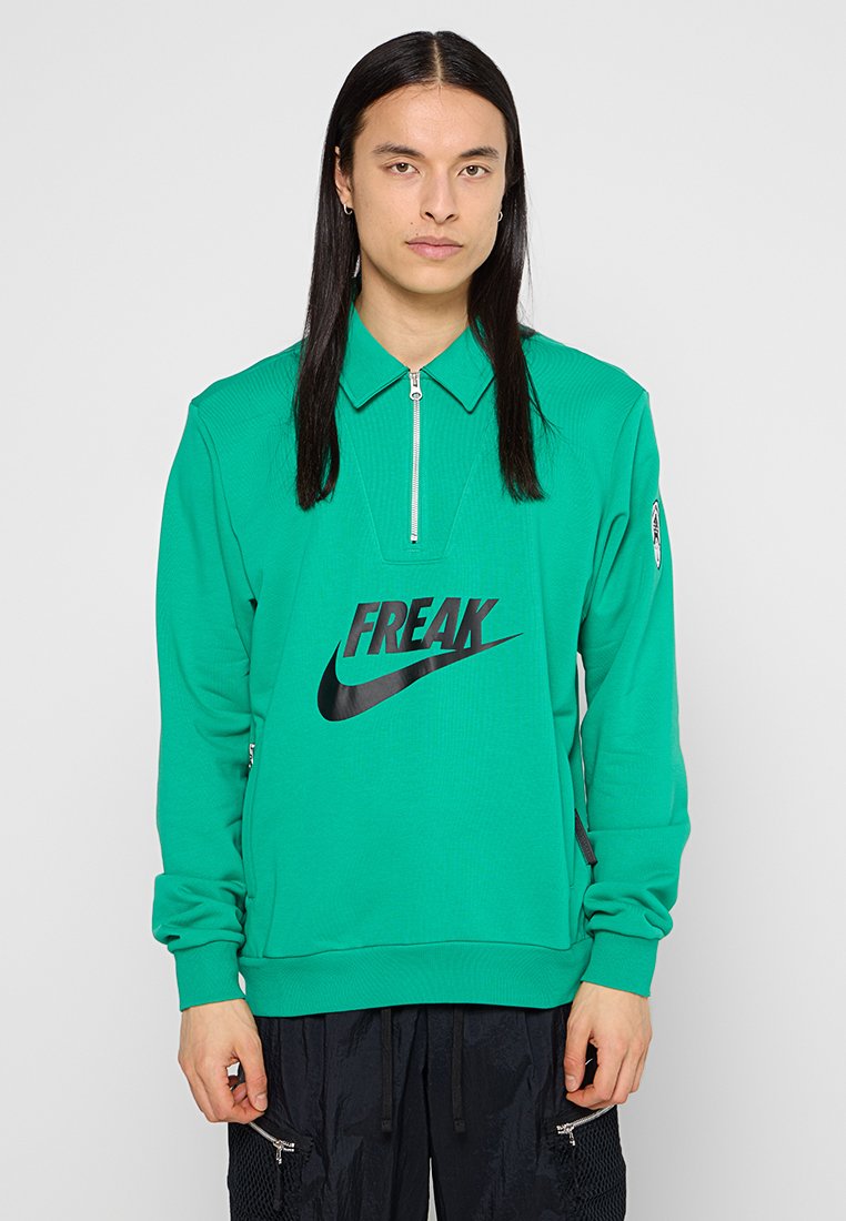 Junge Person mit langen schwarzen Haaren, die einen grünen Zip-Up-T sweatshirt mit "FREAK" und dem Nike-Logo sowie schwarze Hosen mit Reißverschluss-Taschen trägt.