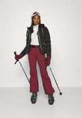 Schwarze Pufferjacke, weißes Oberteil, burgunderfarbene Skihose, schwarze Handschuhe. Skistöcke in der Hand, mit Skibrille. Schwarze Skischuhe mit Riemen.