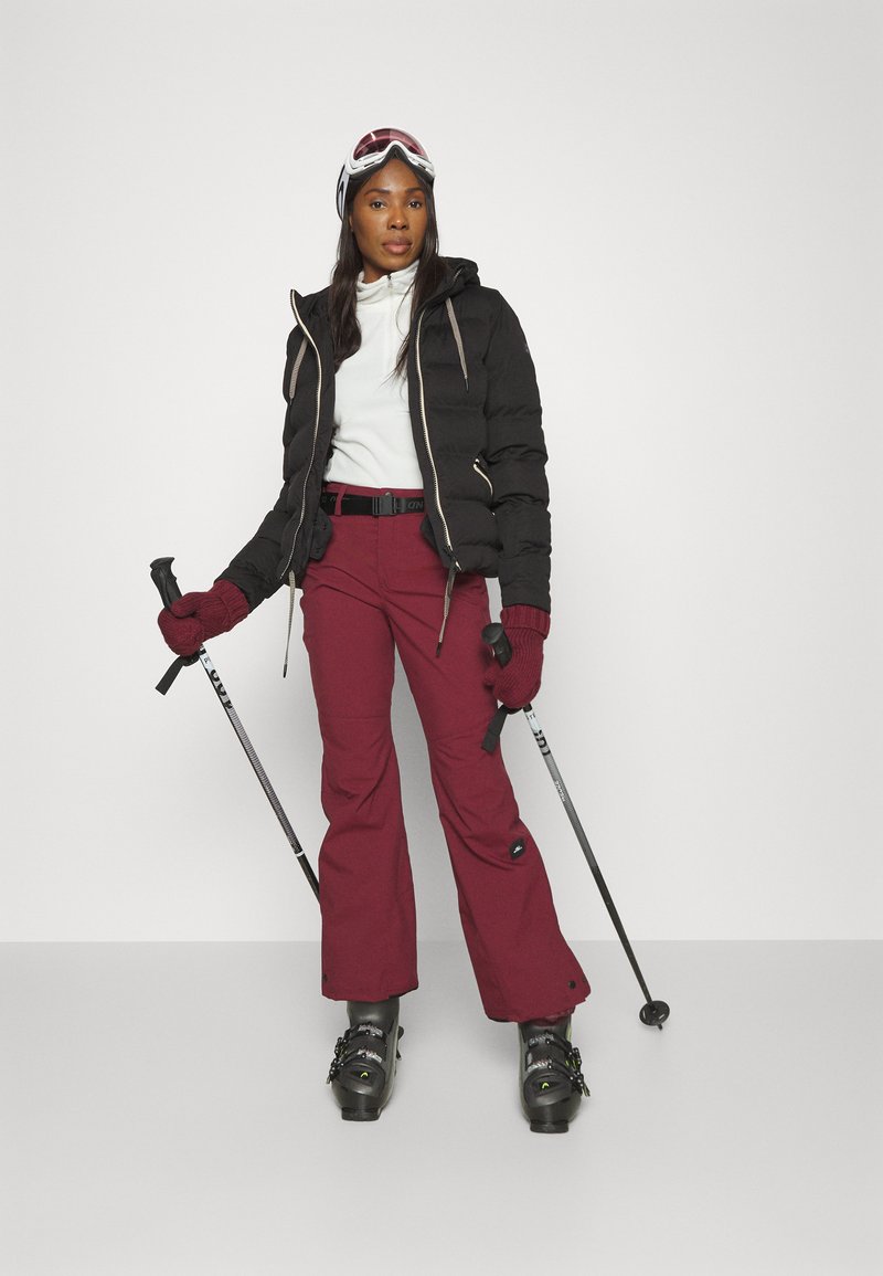 Schwarze Pufferjacke, weißes Oberteil, burgunderfarbene Skihose, schwarze Handschuhe. Skistöcke in der Hand, mit Skibrille. Schwarze Skischuhe mit Riemen.