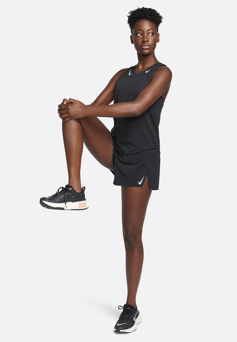 Schwarzes sportliches Tanktop mit durchbrochenem Design, kombiniert mit schwarzen Shorts. Getragen mit schwarzen Laufschuhen mit orangen Akzenten.