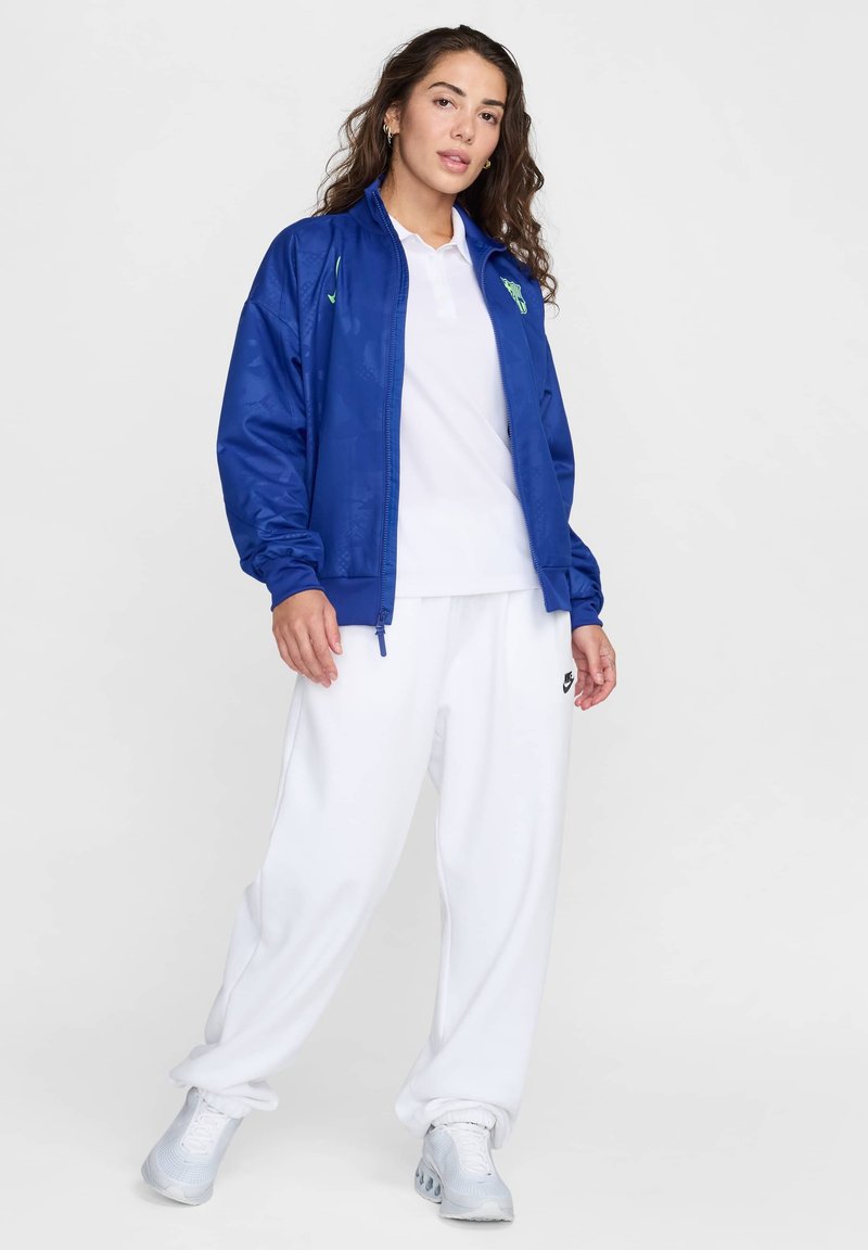 Blaue Zip-Jacke, weißes Polohemd, weiße Jogginghose, graue Turnschuhe. Die Jacke hat ein Logo und gerippte Bündchen; die Hose ist weit geschnitten.