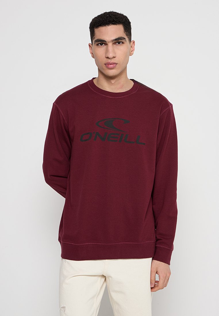 Junger Mann in einem burgunderfarbenen O'Neill-Sweatshirt und hellen Hosen, der vor einem einfarbigen hellgrauen Hintergrund steht.